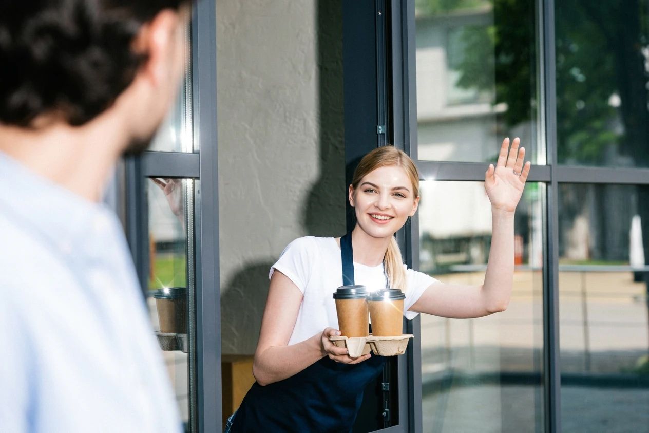 A barista waving to a regular as she brings out two coffees